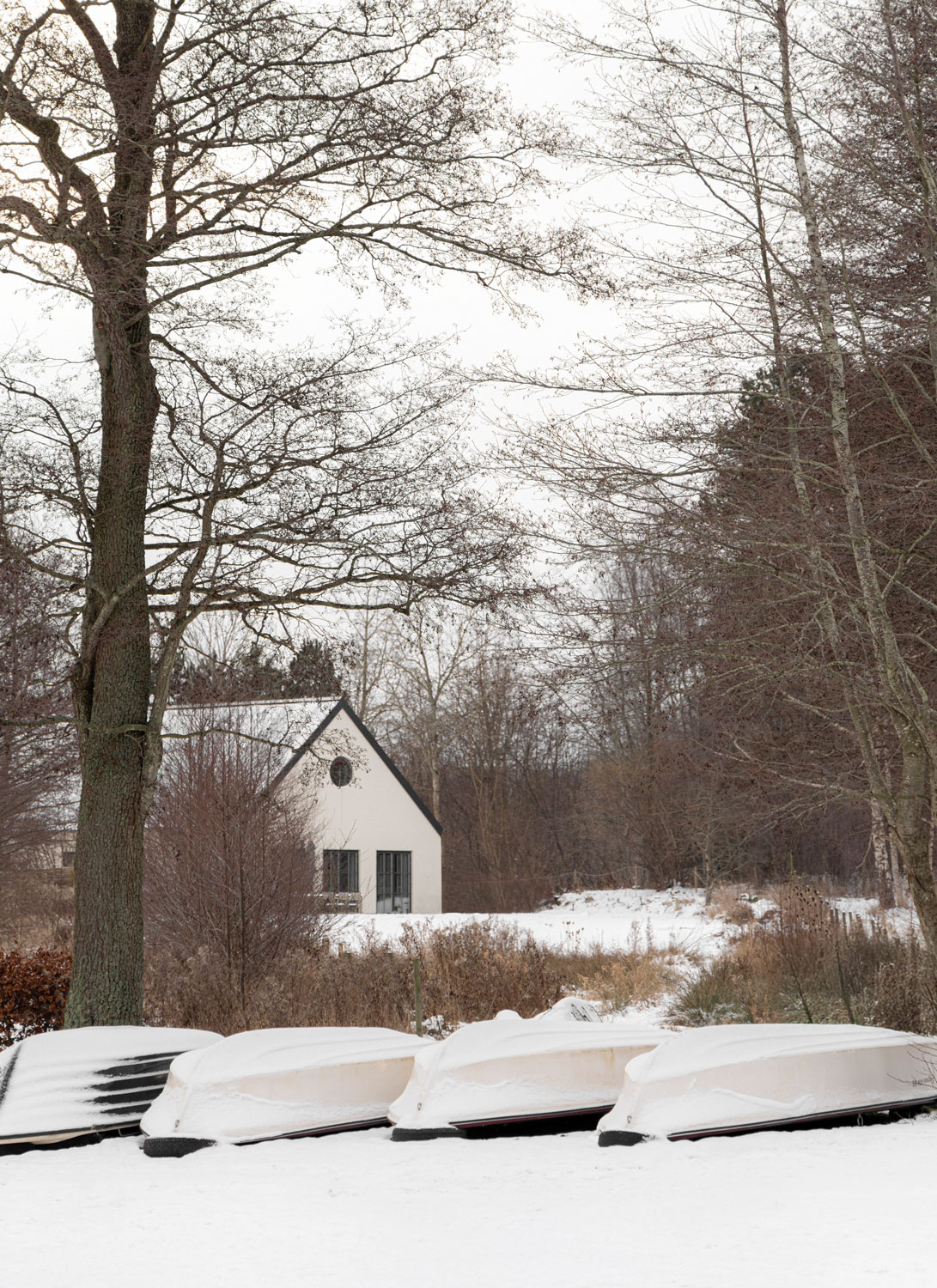 Snow-covered lake house site with trees, stored boats, and a small cabin near the water